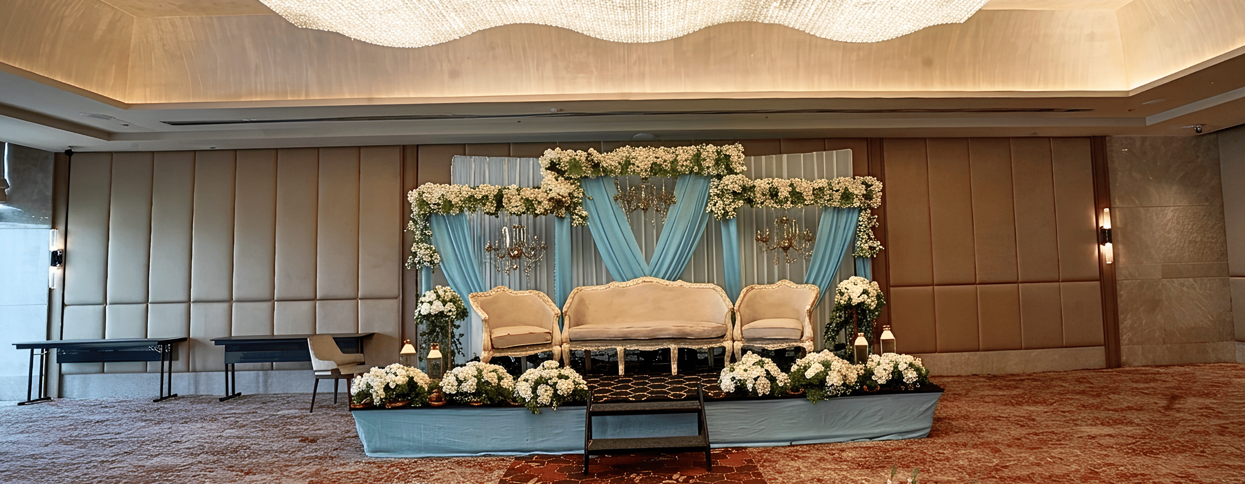 A stage set up for an event inside a hall, featuring seating, drapes, floral arrangements, a carpeted floor, and a chandelier.