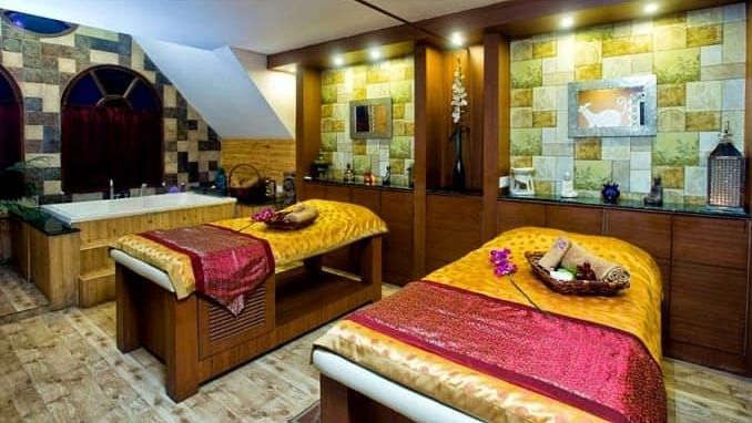 A cosy spa treatment room at Hotel Mount View featuring two massage beds adorned with red and yellow throws. The room features warm lighting, wood accents, and shelves displaying various spa products and decorative items.