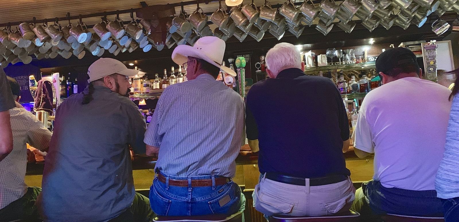 A dimly lit bar interior shows four people sitting on stools with their backs to the viewer. Above them, glasses hang neatly, and the wall features framed items. - Brewery Gulch Inn & Spa.