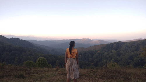 A person stands on a ridge looking at distant mountain ranges at dusk.