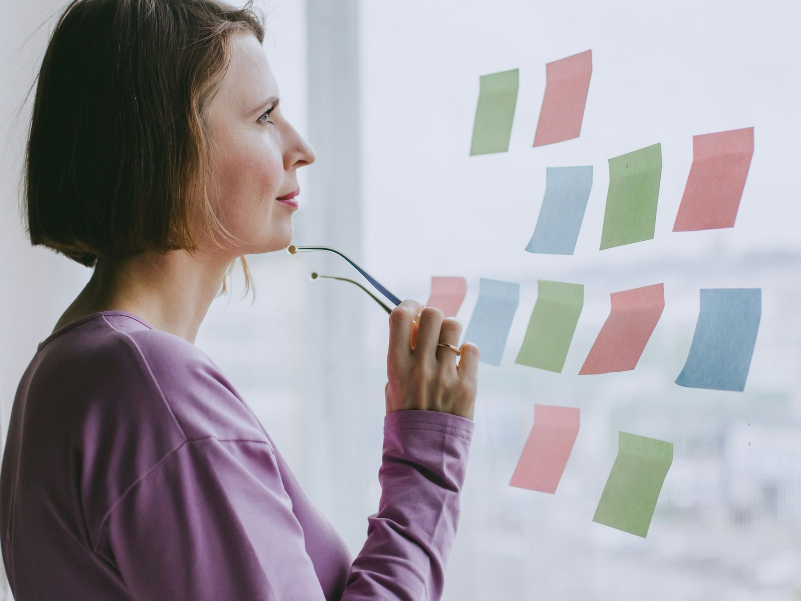 A woman in a purple top looks thoughtfully at a window covered with colorful sticky notes, holding glasses to her chin.