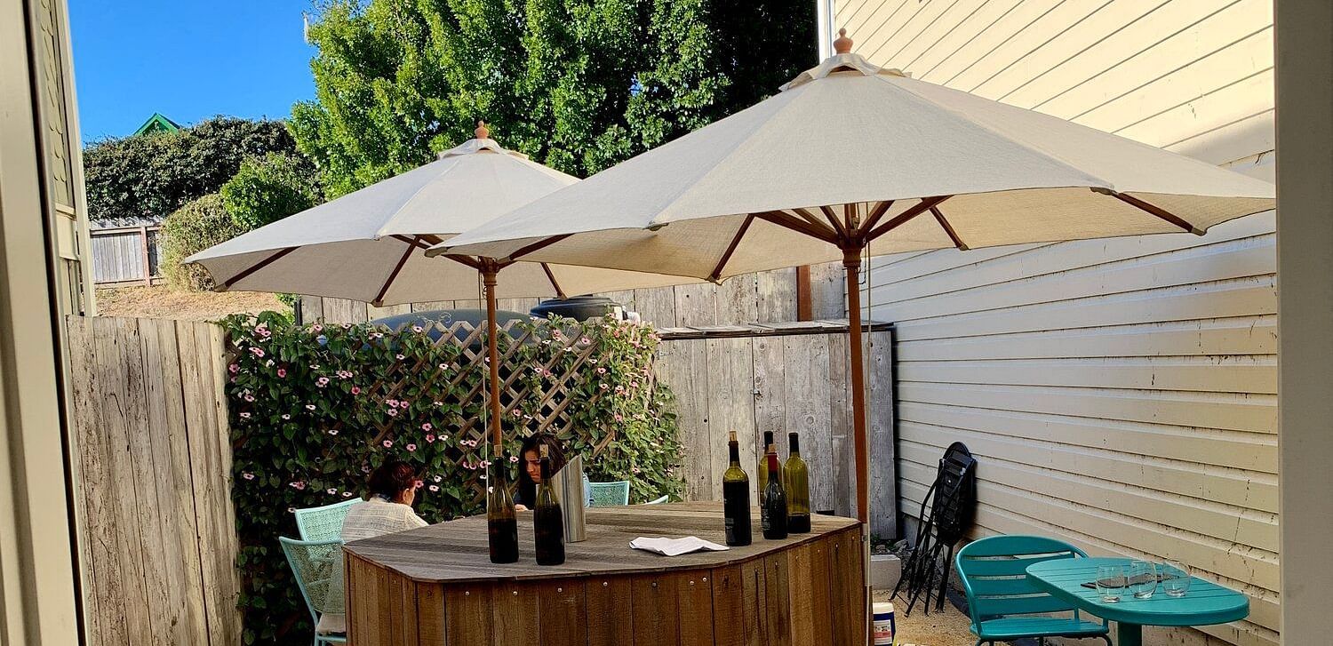 Seen through a doorway, an outdoor patio features a wooden bar with wine bottles, umbrellas, and colorful chairs.