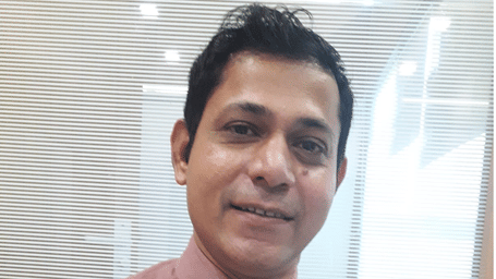 A smiling man in a pink shirt, standing in front of a glass partition with white horizontal lines.