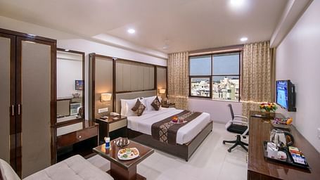 A practical hotel room contains two single beds, a dedicated study desk, a television for guests, and a window showing a cityscape.