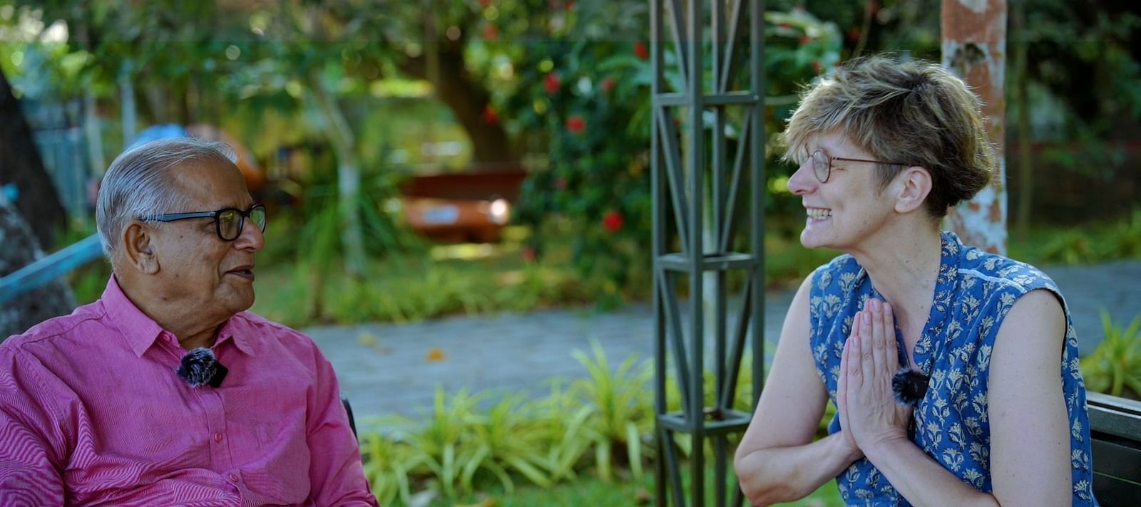 Two people sit facing each other outdoors, engaged in conversation, with greenery in the background.