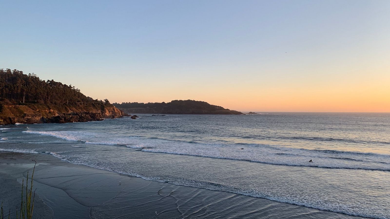 A coastal view with a beach, ocean waves, and a headland in the distance under a clear sky at sunset.
