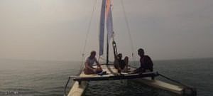 A small sailboat with multiple people aboard sails on calm water under a bright, hazy sky.