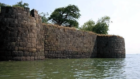 A fort wall standing along the edge of water with greenery visible on the top.