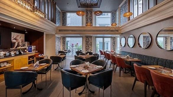 A side-angled view of a restaurant seating area with tables and chairs, warm lighting and windows at Hotel Saket 27, New Delhi