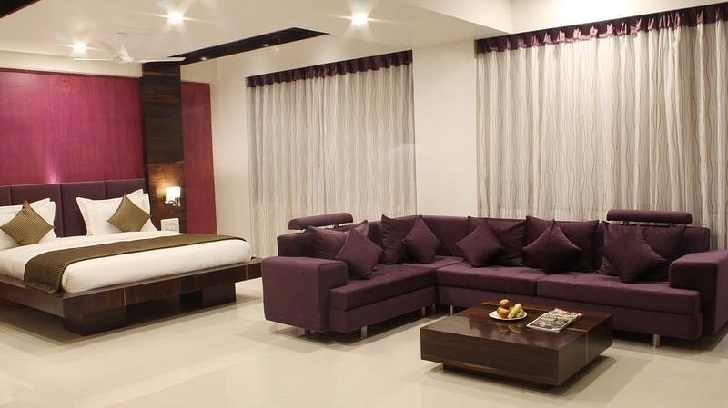 A spacious hotel suite features a king size bed, a comfortable brown sofa set around a coffee table, and large windows with sheer curtains.
