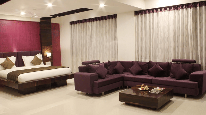 A spacious hotel suite features a king size bed, a comfortable brown sofa set around a coffee table, and large windows with sheer curtains.