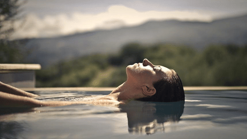 A woman floats peacefully in an outdoor pool, eyes closed, surrounded by nature and distant mountains under a soft, calming sky. 