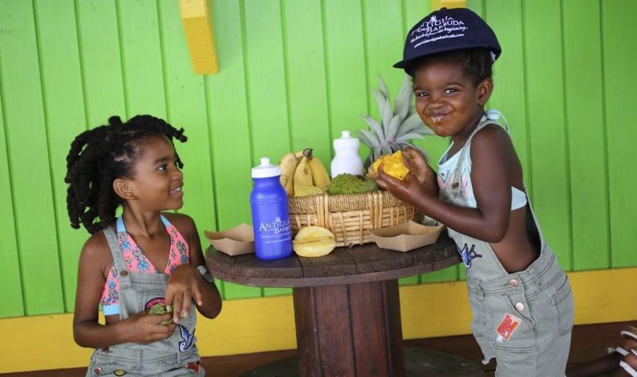 Two children are seated at a small table outdoors, one holding a drink and the other holding fruit.