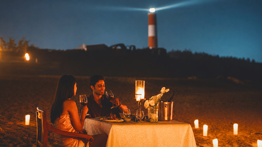 Couple enjoying a private candlelit beach dinner at night with a beacon lighthouse glowing in the background at MAYFAIR Palm Beach Resort, Gopalpur-on-Sea. 