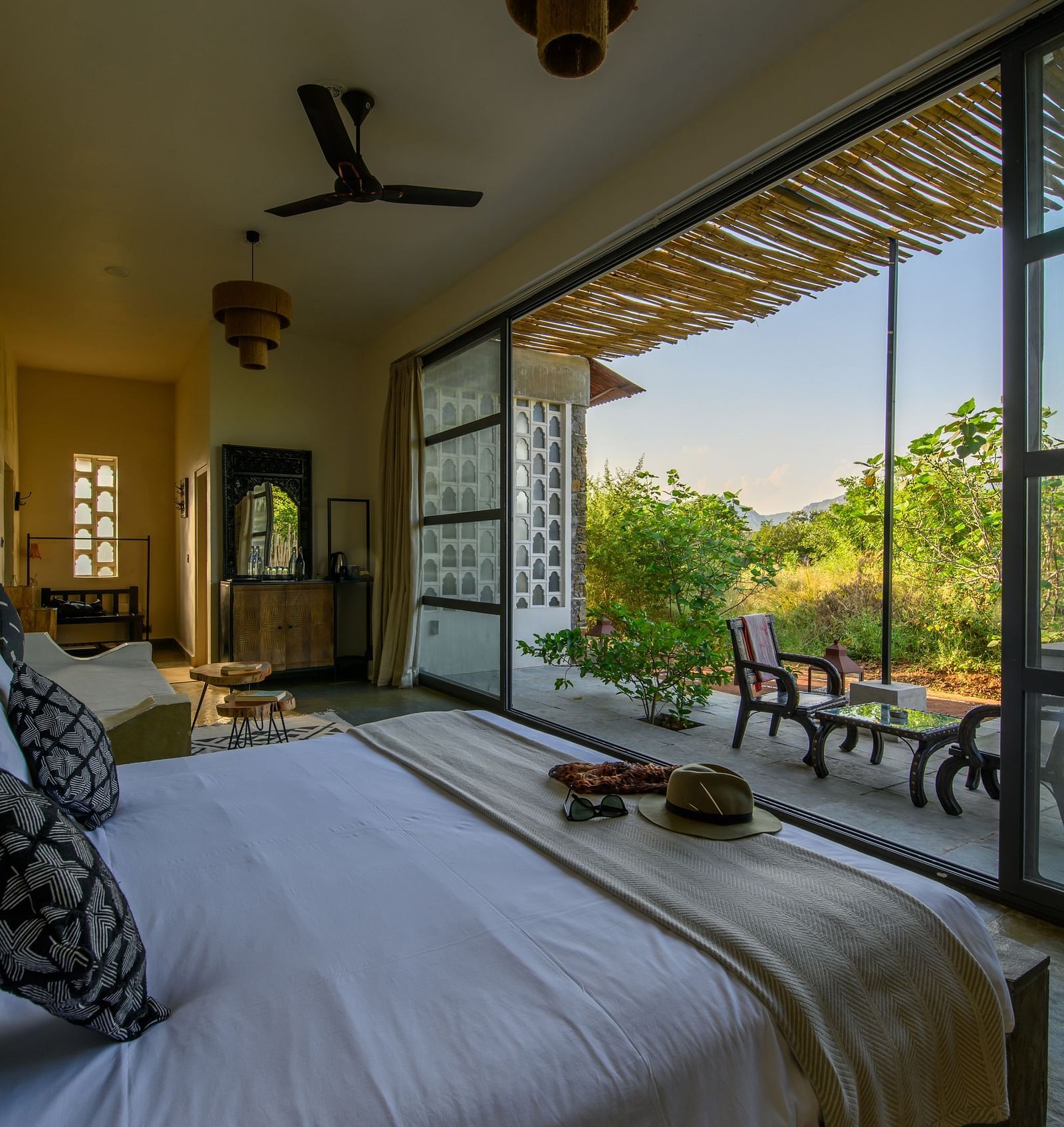An image of a room with a queen size bed, chandelier, ceiling fan, other decorative items and an expansive glass door leading out - Utsav Camp Sariska