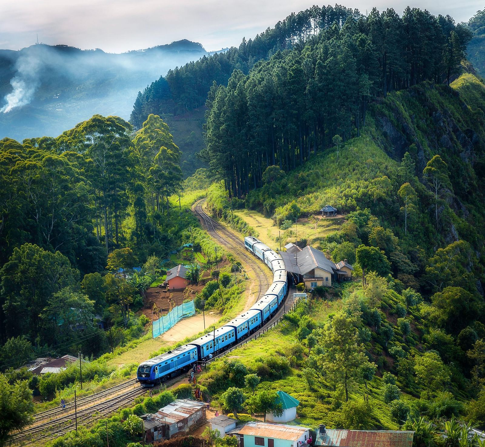 Train going through raintracks with houses and greenery on both sides and mountains along with pine forest in the distance, imitating sceniery of delhi to joshimath train journey