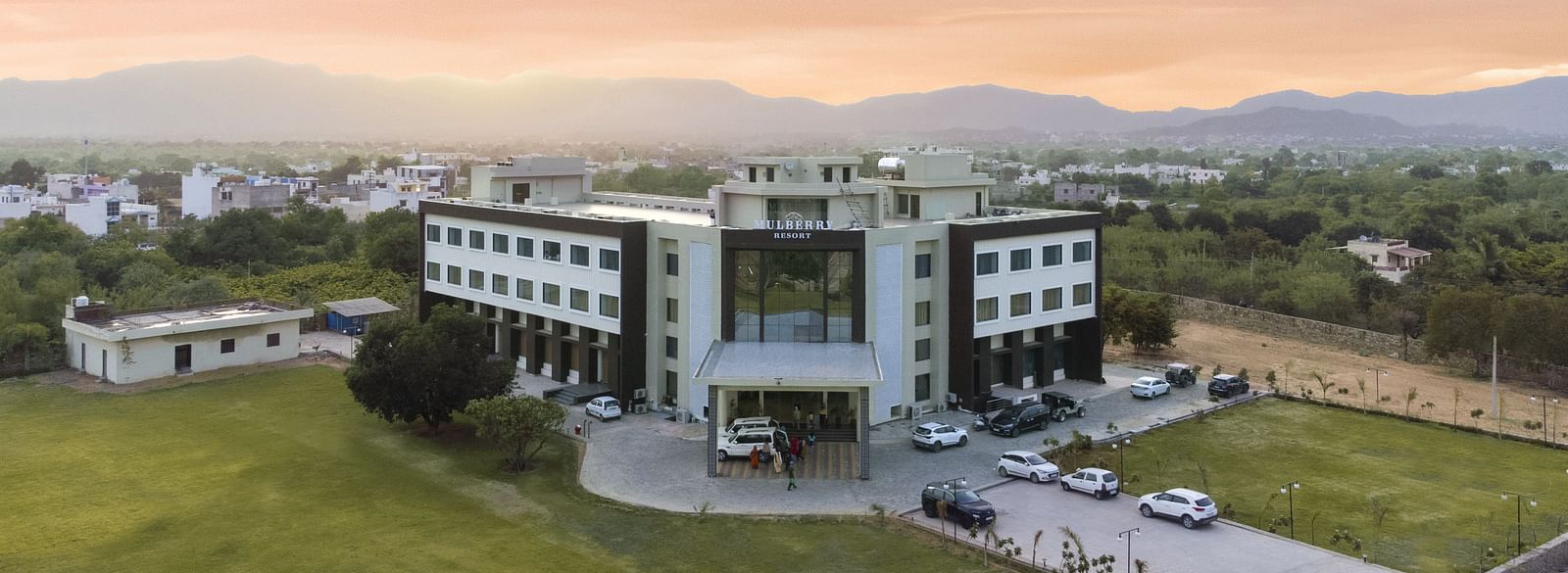 An aerial view of the Bamboo Saa Mulberry Resort, Pushkar at sunset or sunrise surrounded by green lawn and the backdrop of hills.