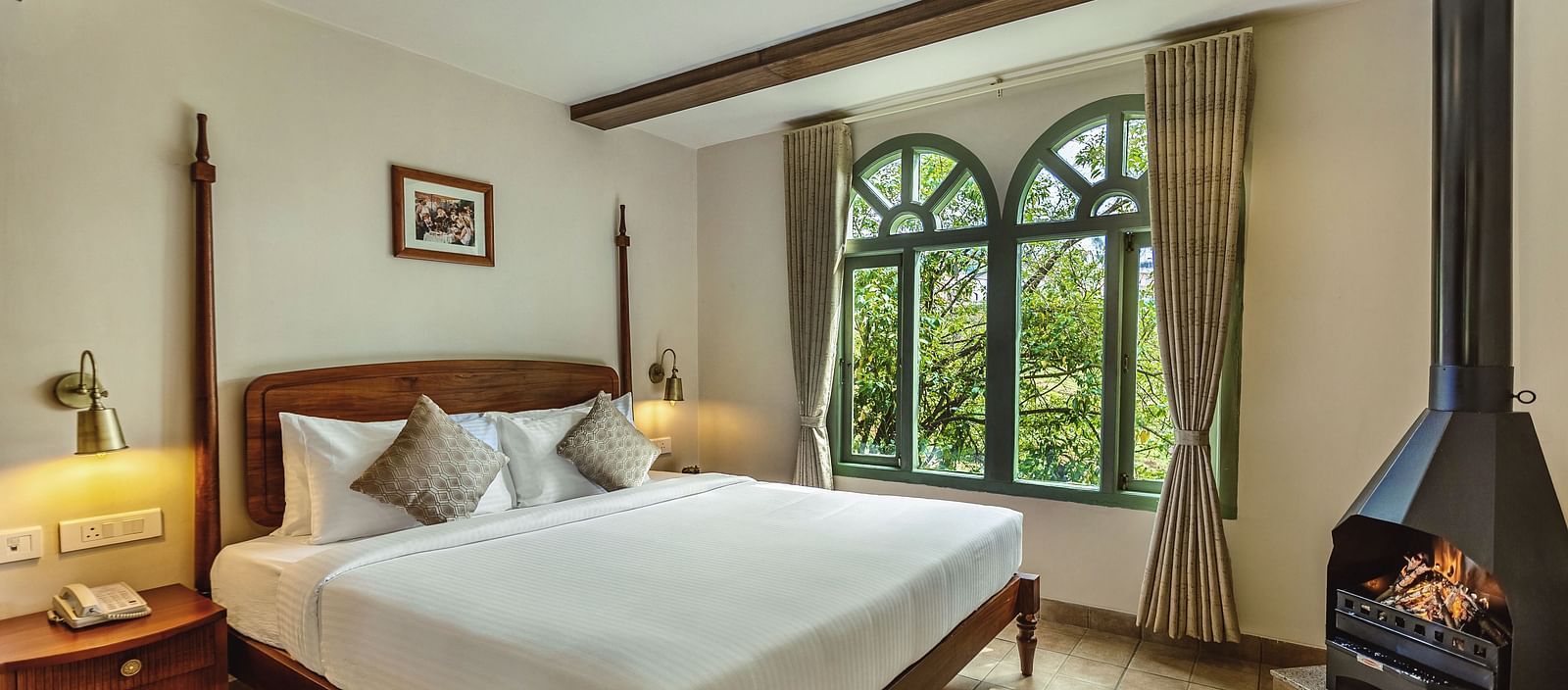 Sunlit bedroom at Mango Hill Shola, Ooty with a wooden bed, side tables, and large arched windows offering lush green views.