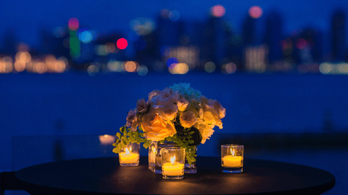 many candles lit and kept on a table around a vase with flowers and a backdrop of the city skyline