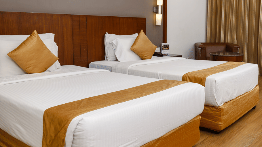 Twin bed room at Featherlite Evoma Hotel with beds dressed in white and gold linen, wooden headboards and warm ambient lighting nearby.