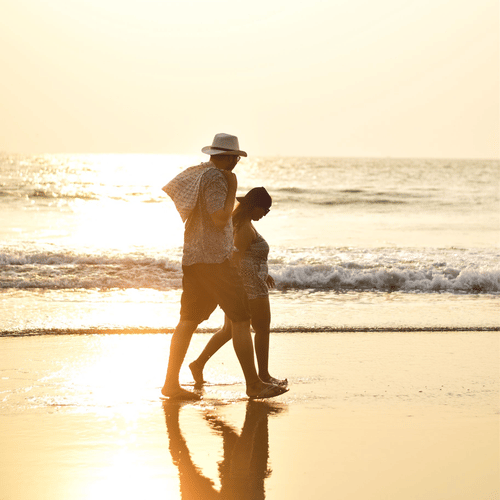 A couple walking on a wet shoreline of a beach with a bright sun reflecting the rays on the ground and the ocean.