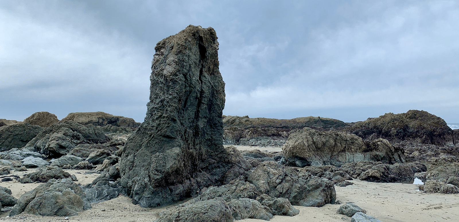An image of beach side featuring several rocks and there is also one huge rock upright