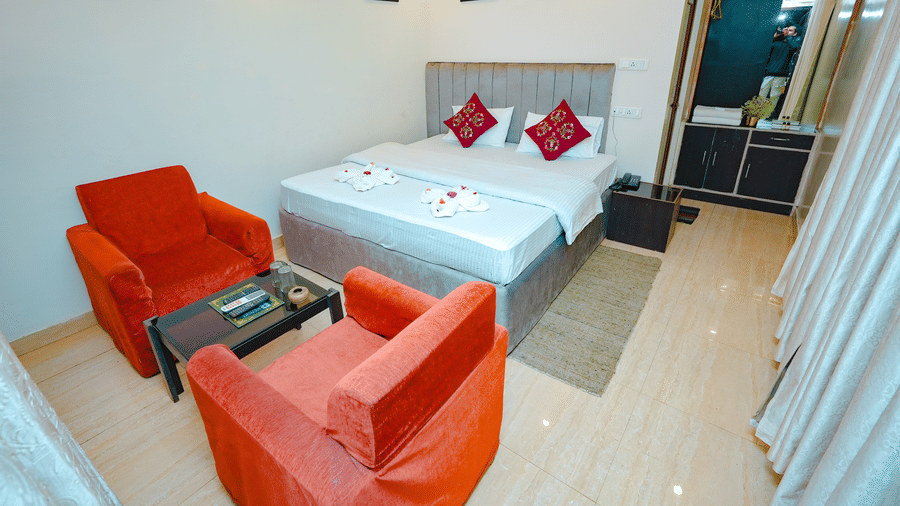 A spacious hotel bedroom featuring a double bed, white bedding with red accents, and a seating area with two orange armchairs.