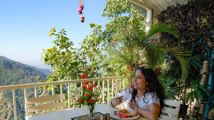 A lady sitting at a rooftop restaurant with plants in the background, enjoying her meal while taking in the scenic mountain view near the Ganga River.