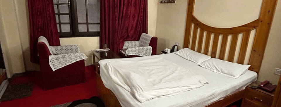 View of a bed with white linen, a wooden headboard, red carpet, and a black and white patterned rug at Hotel Dolphin Darjeeling.