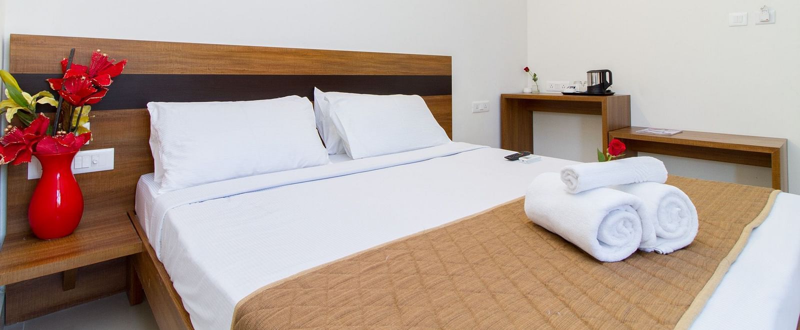 A room presenting a bed, neatly rolled white towels, bedside tables and a red vase placed on it at Sanctum Suites Whitefield, Bangalore.