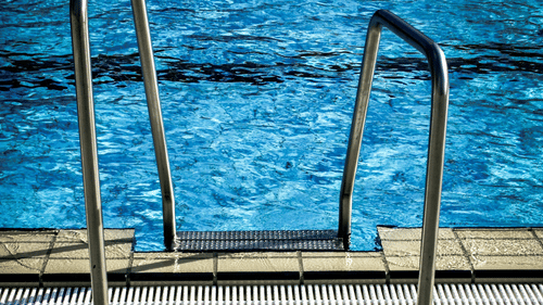 The edge of an outdoor swimming pool, showing metal steps for entry and exit, against a surrounding pattern of paved tiles.