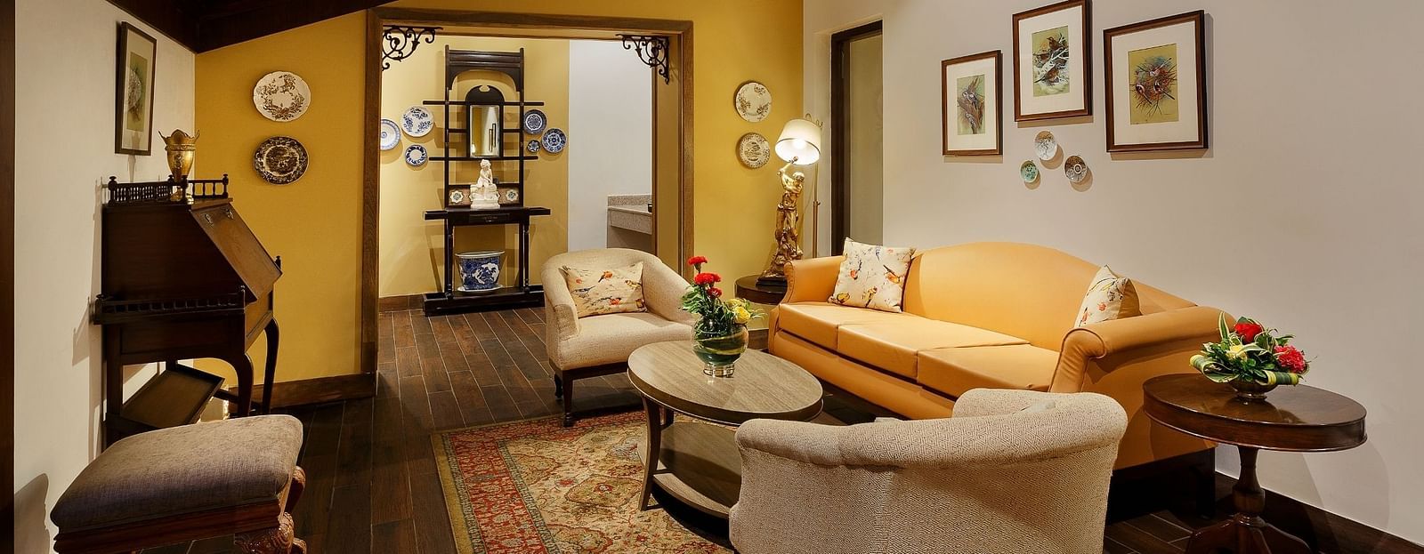 An image of the living area with Sofas that could seat 5 people, spaciously arranged furniture, multiple wall paintings, and a slanted wooden ceiling which accentuates the ambience.