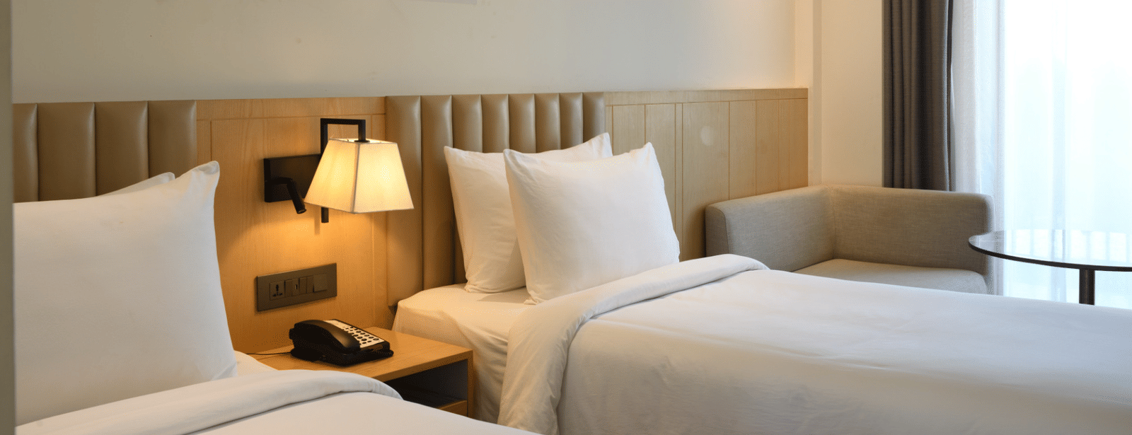 Neatly arranged twin-bed room with white bedding, a classic table lamp, wooden headboard, sheer curtained windows, and wall art above.