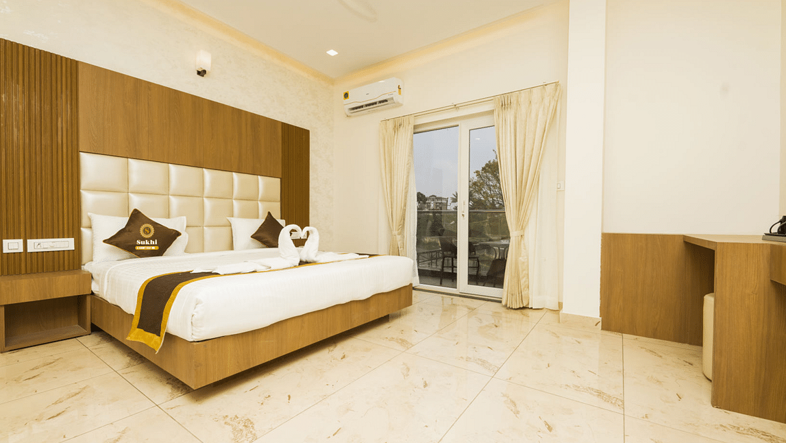 Super Deluxe Room With Balcony featuring a king size bed with a wooden headboard, and decorative pillows, alongside light-coloured walls, tiled flooring.- Sukhi - A Luxury Stay Inn, Chikkamagaluru