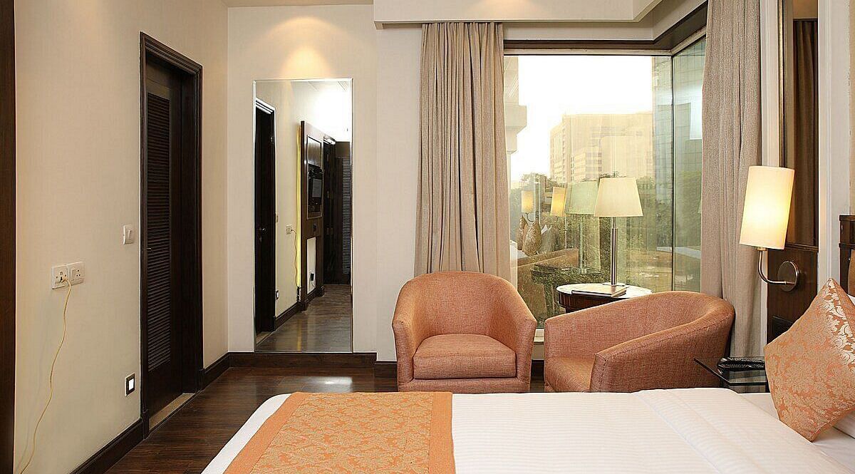 Executive Room with a double bed, bedside table with a lamp on top, cushioned seating with a table in front, and a window behind at Inde Signature Tower, Gurgaon
