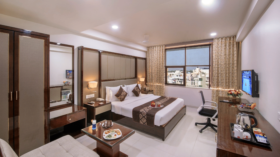 A practical hotel room contains two single beds, a dedicated study desk, a television for guests, and a window showing a cityscape.
