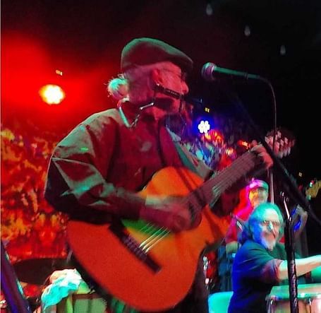 A musician from the Pura Vida Band performing on stage, playing a guitar under colorful lights, with the band members in the background.
