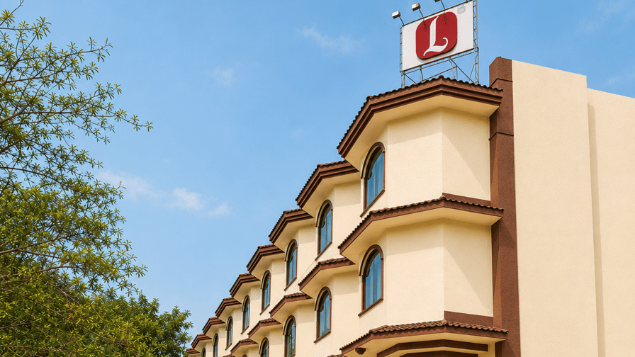 Exterior view of a multi-story cream-coloured hotel building with a red rooftop Lords logo sign, paved driveway, and a parked silver car in front - Lords Inn Vadodara