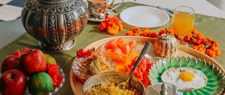A breakfast spread laid out on a table on a balcony, featuring fruits, eggs, drinks, and other dishes, with a view of blue railings and greenery | Dileep Kothi, Jaipur