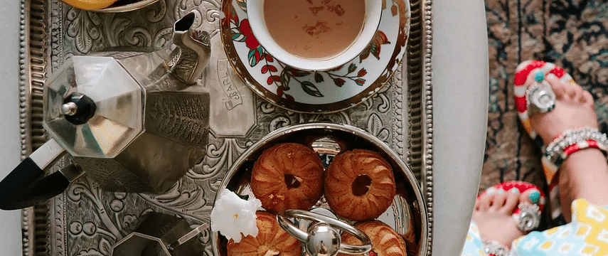 A flat lay of a silver tray with coffee, pastries, and a yellow flower on a white table, with a person's patterned dress and feet visible at the edge | Dileep Kothi, Jaipur