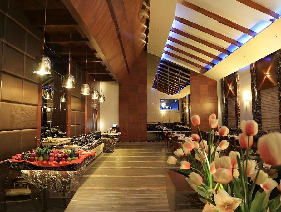 A view of the Aroma restaurant in Dhanalakshmi Srinivasan Hotels, Perambalur, featuring a buffet counter on the left and dining tables in the background. The restaurant has wooden ceiling beams and a floral arrangement in the foreground.