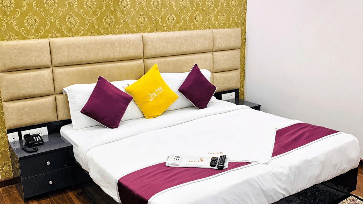 Bedroom of Lime Tree Hotels & Service Apartment Private Limited featuring a padded headboard, pink and yellow cushions, and a patterned rug.