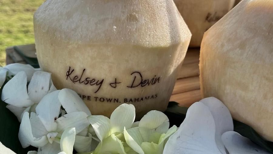 Coconuts with straws dipped in are placed on a bamboo table near the beach side. The coconuts have carvings with a couple's name Kelsey and Devin and are placed with orchid flowers. 