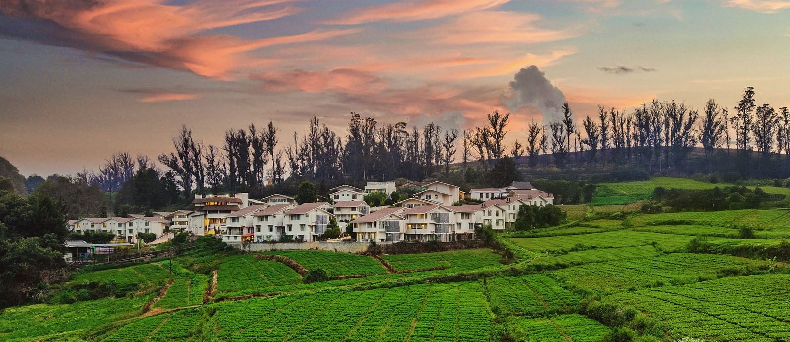 Lush green fields with a backdrop of white cottages nestled under a vibrant sunset sky in a hilly landscape.