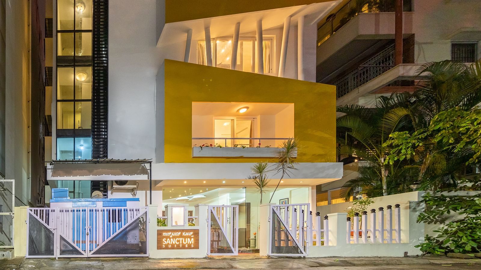 A facade of Sanctum Suites Domlur, Bangalore with warm lighting and view of balconies shot at night.