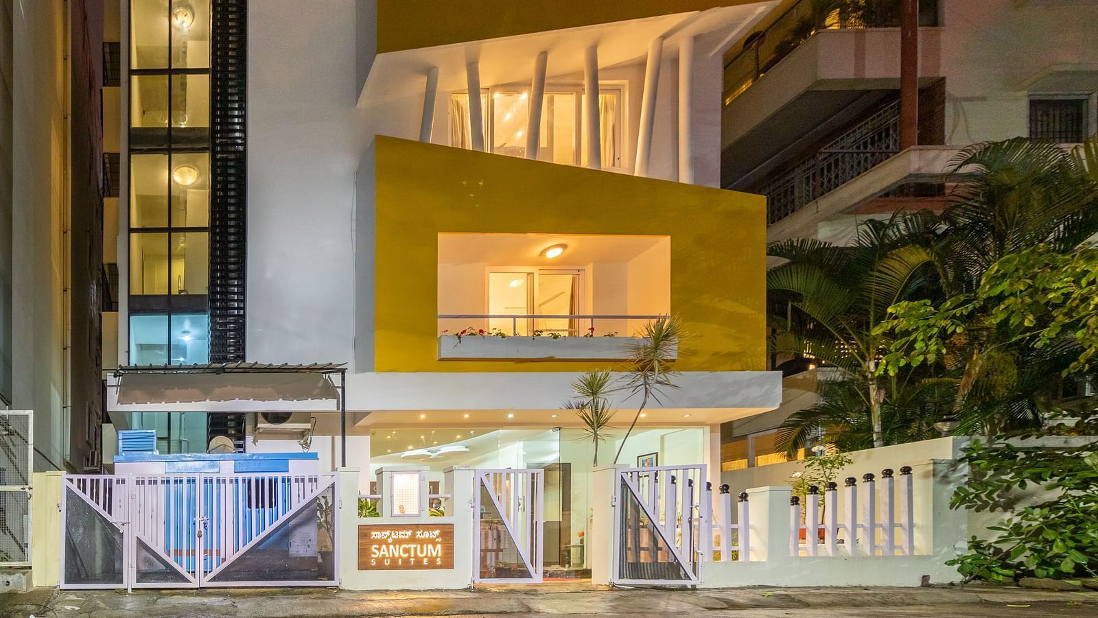A facade of Sanctum Suites Domlur, Bangalore with warm lighting and view of balconies shot at night.