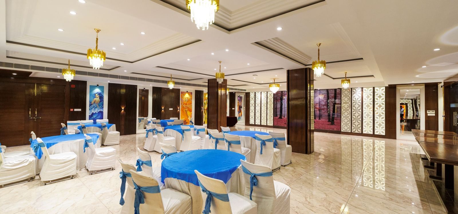 Banquet hall at Inde Reva Resorts Rishhikesh set up with round tables, white chairs, blue covers, and ceiling chandeliers.