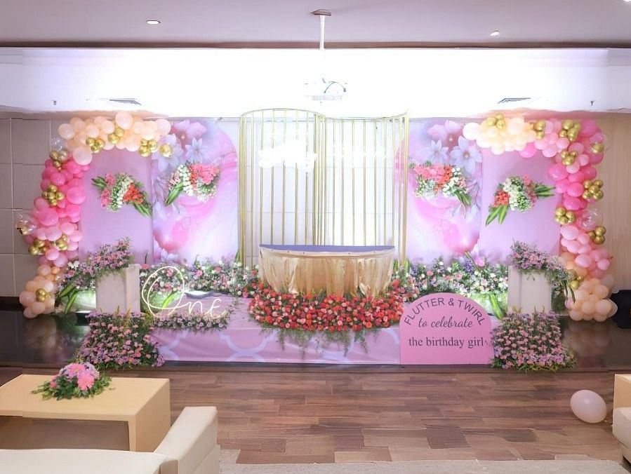 A decorated hall in Dhanalakshmi Srinivasan Hotels, Perambalur, set up for a celebration with a stage adorned with pink and gold balloons, flowers, and a banner. There are sofas and a coffee table in the foreground.