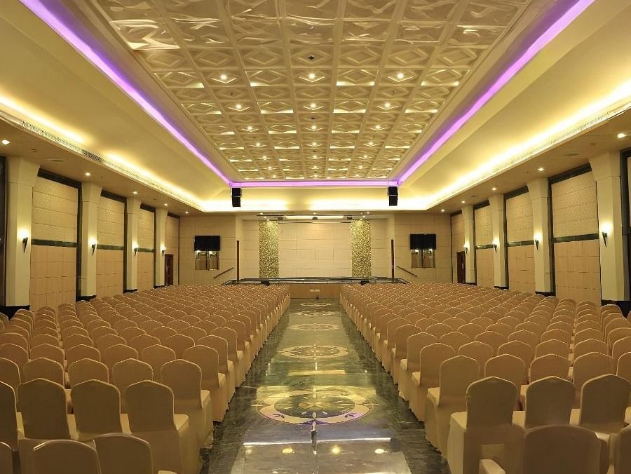 A large empty hall in Dhanalakshmi Srinivasan Hotels, Perambalur, with rows of chairs facing a stage area. The hall has dark marble flooring and a patterned ceiling with lighting.