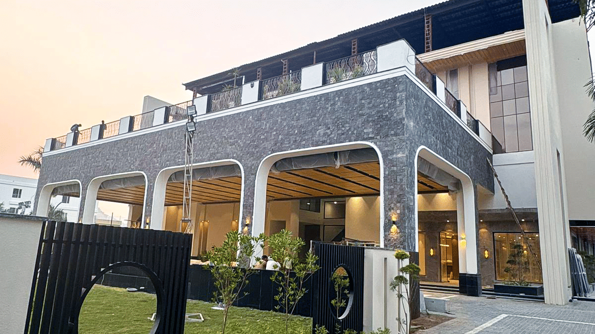 A daytime view of the modern facade of Pride Premier Alwar, featuring dark stone cladding, large windows, and manicured landscaping near the entrance.